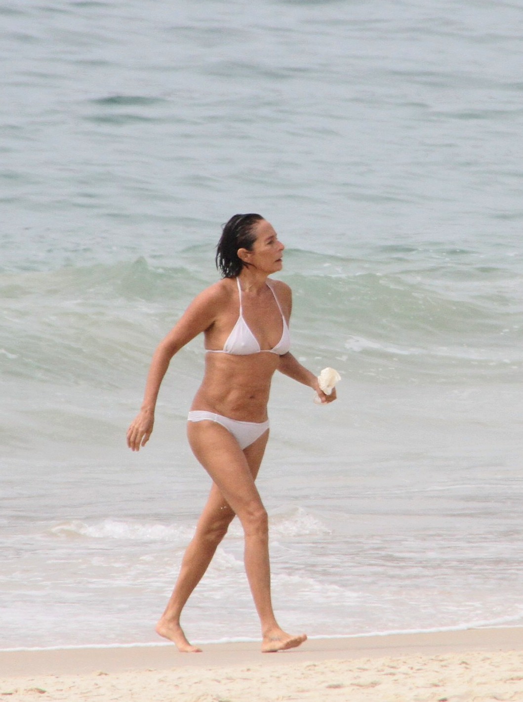 Mulher branca, de cabelo curto, usando um biquíni branco e saindo do mar segurando um pacote branco