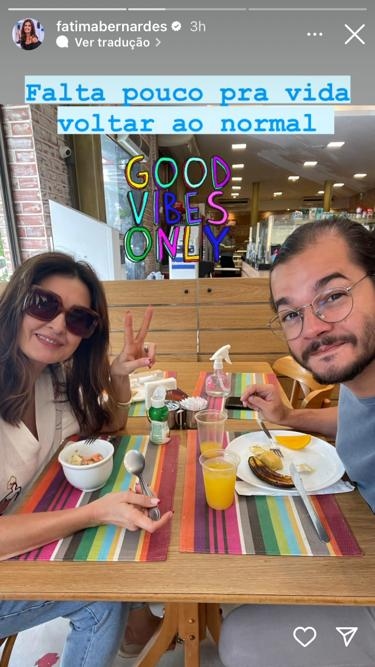 A apresentadora Fátima Bernardes tomando café da manhã ao lado no namorado, o deputado federal Túlio Gadêlha. Ela usa óculos de sol quadrados, uma blusa branca e uma calça jeans, e ele veste óculos de grau redondos, uma camiesta azul e uma calça cinza claro. Sobre a mesa de madeira, há uma salada de frutas, dois copos de suco de laranja e um prato com frutas