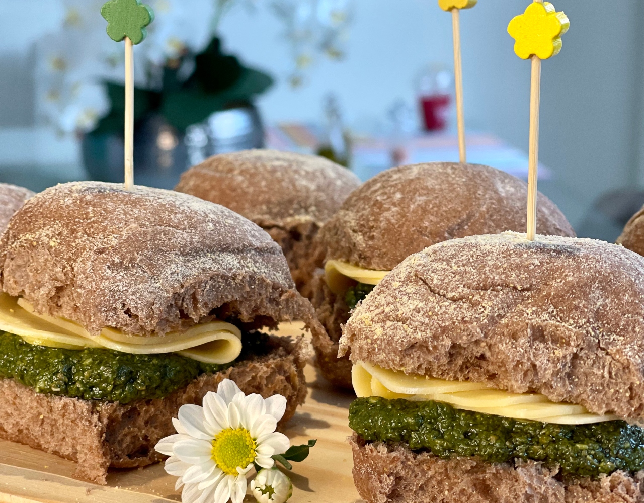 Sanduíches de queijo prato com molho pesto servidos com palitos de madeira, com flores verdes e amarelas na ponta